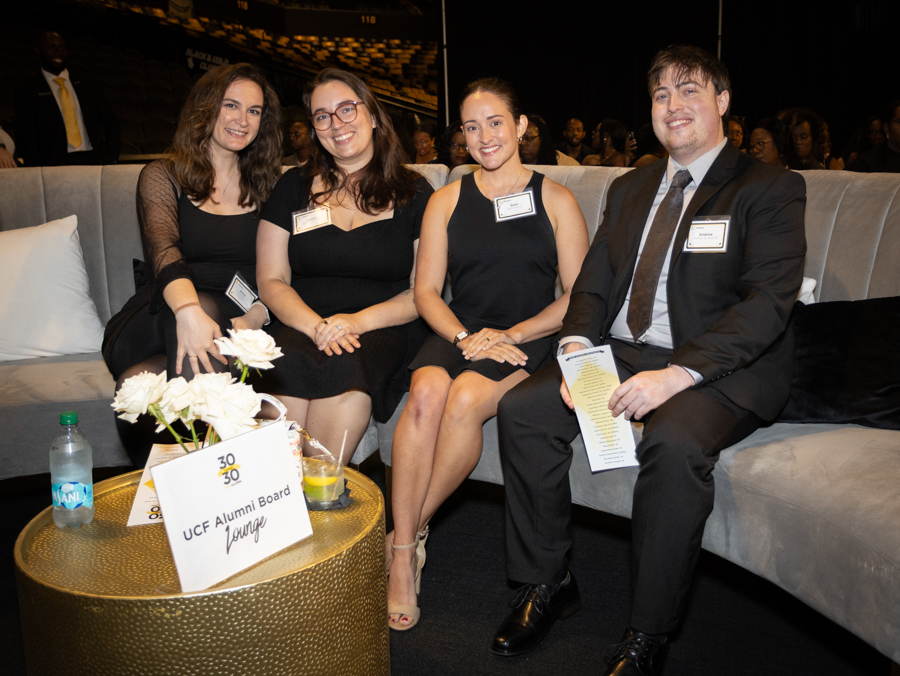 Four people in black attire sit on a gray sofa at a formal event, smiling, with a gold table in front and a sign that reads 'UCF Alumni Board Lounge'.
