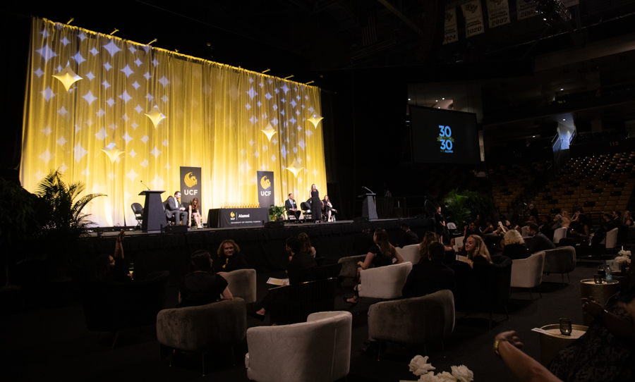 Panel discussion on a stage with a yellow backdrop and UCF banners at an indoor event; audience seated in dim seating area.