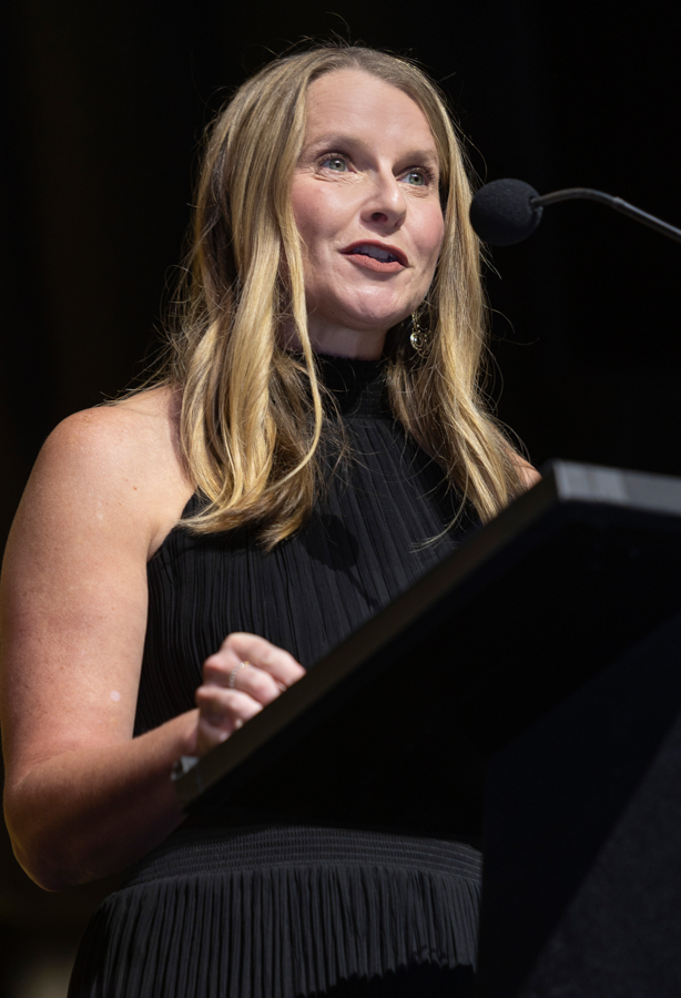 A woman in a black sleeveless dress speaks at a podium with a microphone during a formal event.