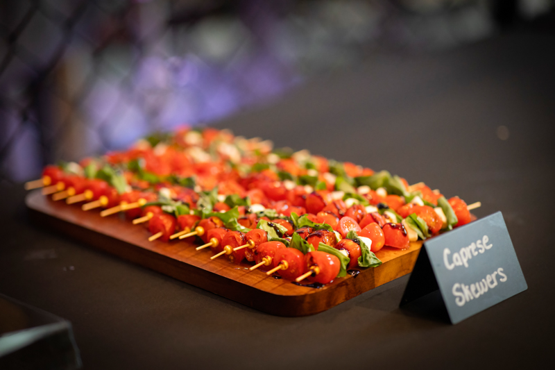 Caprese skewers with cherry tomatoes, mozzarella, and basil arranged on a wooden board at a catering table, with a sign that reads 'Caprese Skewers'