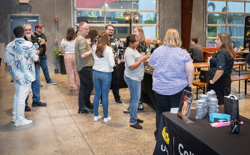 People mingle and chat around a central table at an indoor social or networking event, holding drinks in a spacious venue with exposed floors and a staffed display table nearby.