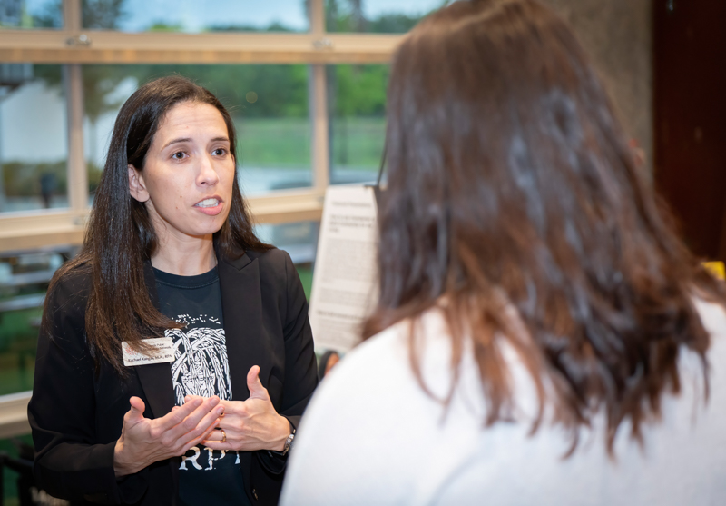 Woman in a blazer explains something, gesturing with her hands during a conversation in a well-lit indoor space with large windows.