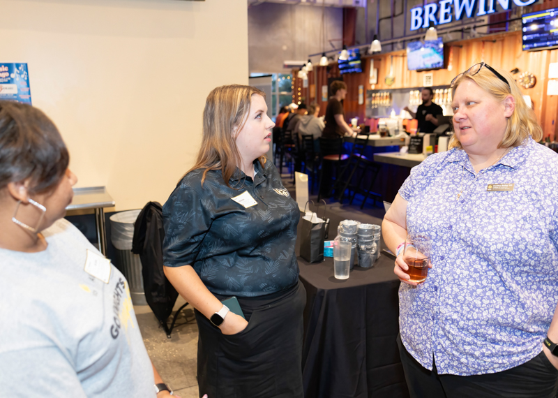 Three women chatting at a brewery event, with a bar and staff in the background.