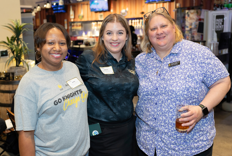 Three women with name tags posing together in a bar or lounge, smiling at the camera; one woman on the right holds a beer glass.