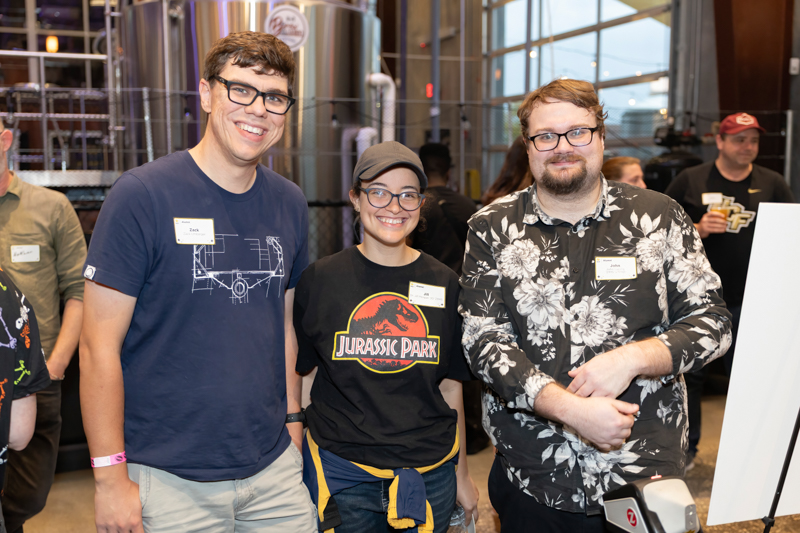 Three smiling attendees with name badges pose together at an indoor event, with metal equipment visible in the background.