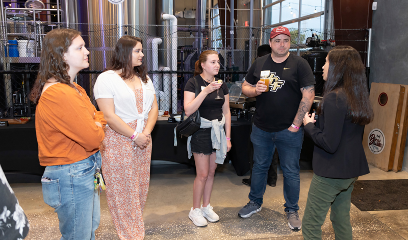 Five people stand in a circle chatting inside a brewery, stainless tanks and equipment visible in the background.