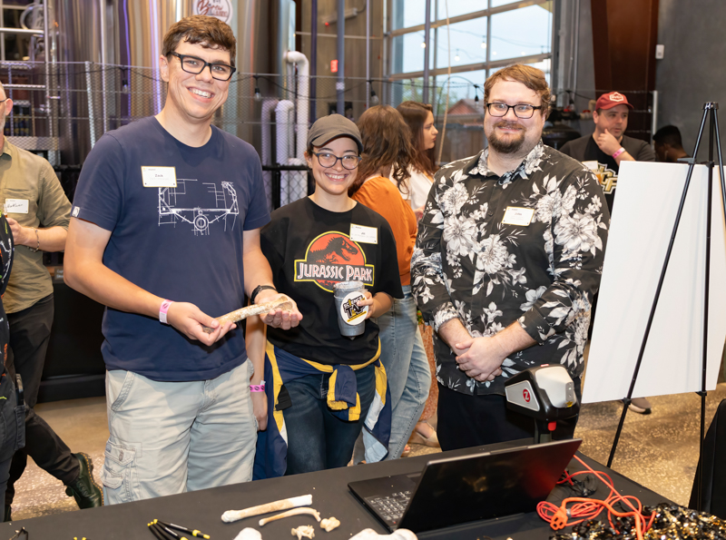 Three people at a convention booth smile for the camera; the left person holds a bone replica while the middle person wears a Jurassic Park shirt and the right person stands near a laptop and whiteboard.