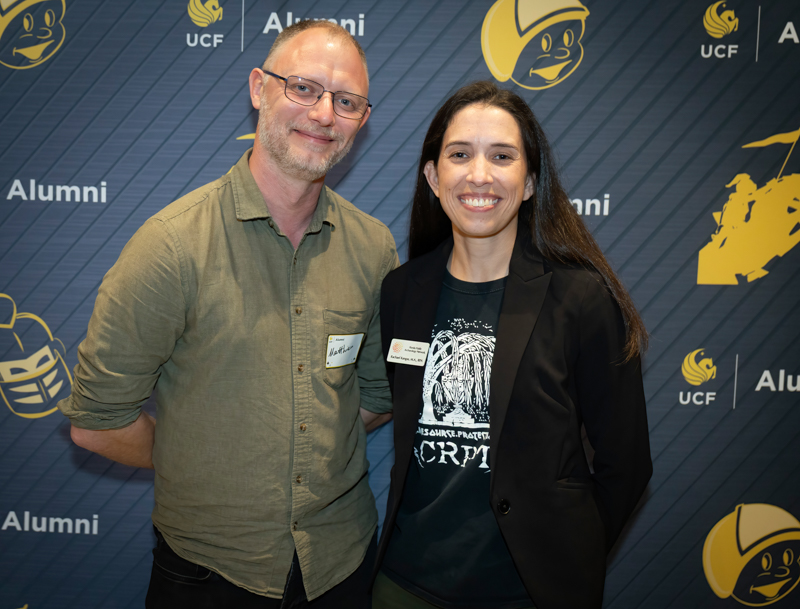 Two adults posing for a photo at an Alumni event, with UCF logos on a blue backdrop.