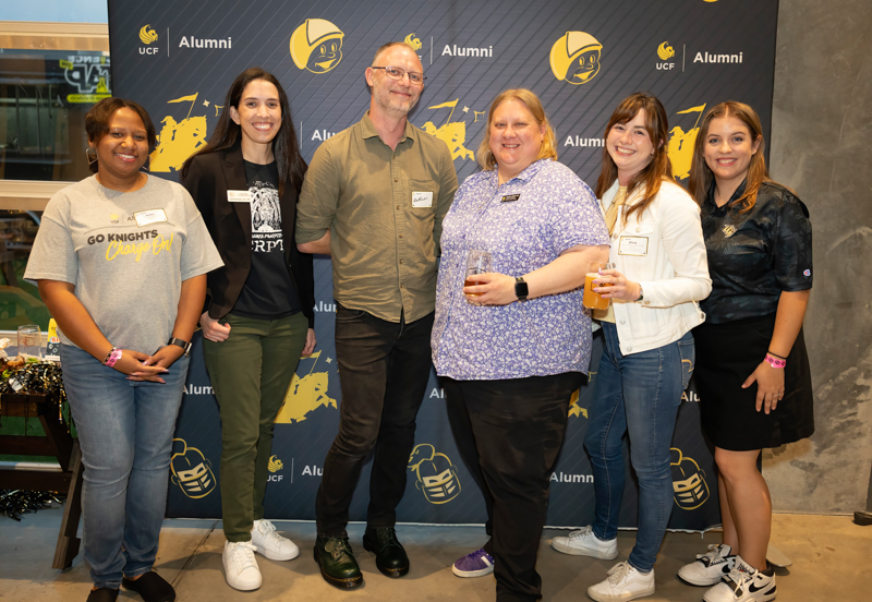 Six adults stand together at a UCF Alumni event, posing for a photo in front of an Alumni backdrop; some hold drinks and wear name tags.