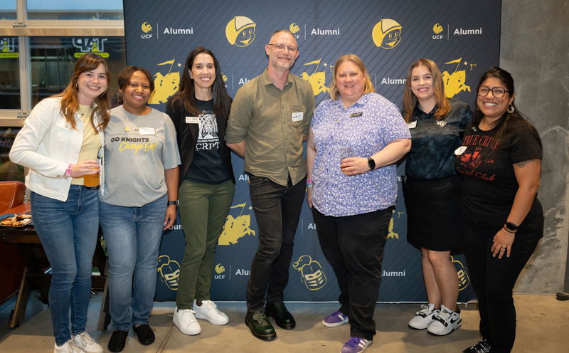 Seven adults posing together in front of a UCF Alumni backdrop at a social event, smiling for the camera.