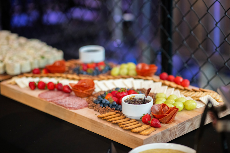 Charcuterie board with cheeses, crackers, fruits, sliced meats, and a dipping sauce on a wooden platter at a party.