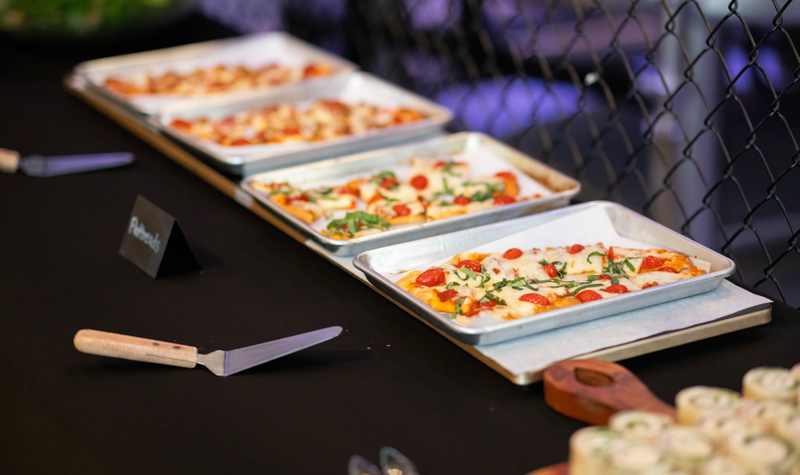 Row of rectangular pizzas with toppings on baking trays at a buffet table.