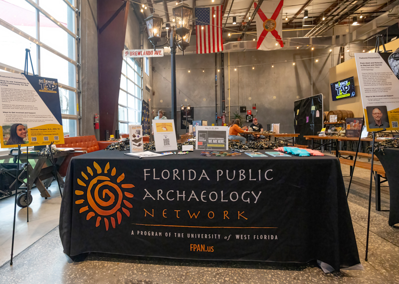Table for Florida Public Archaeology Network at an indoor expo; black cloth with orange sun logo and text, brochures and display boards on the table.