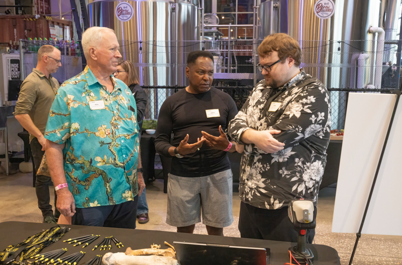 Three men with name tags discuss at a table in a brewery, with stainless tanks in the background and a display of pens and bones on the table.