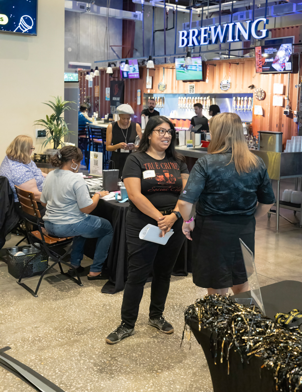 Two women chat at a bustling bar area labeled 'BREWING' with staff and patrons in the background.