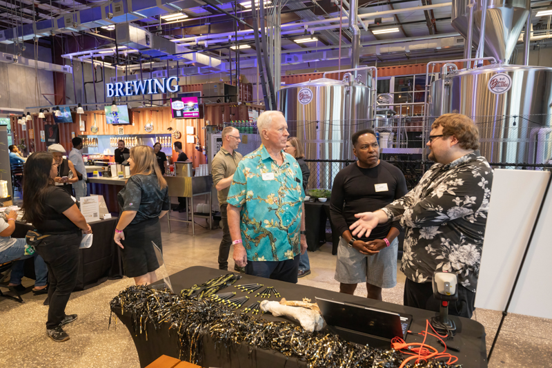 People talking at a brewery tour or open house, with large stainless brewing tanks in the background and a table of items in the foreground.