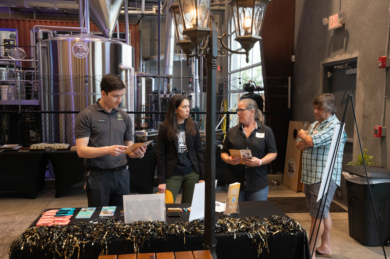 Four people stand around a promotional table at a brewery event, with stainless tanks in the background and brochures on the table.