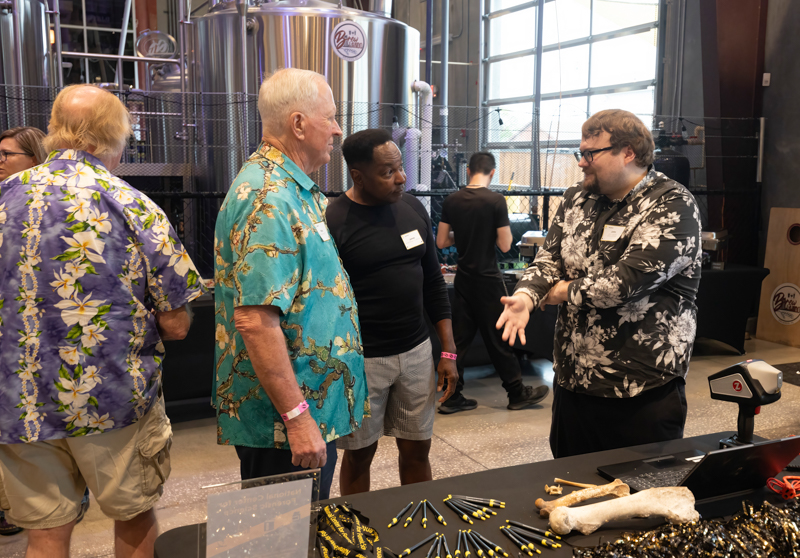 Three men converse at a booth during a trade show, with tools and bone-like objects laid out on the table and large metal tanks behind them.