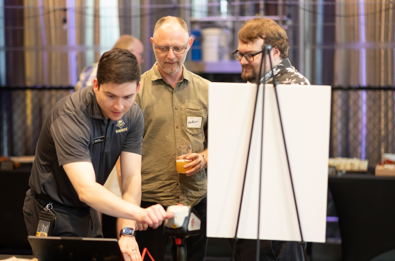 Three men at a tech demo; one leans over a laptop and handheld scanner while two watch nearby.
