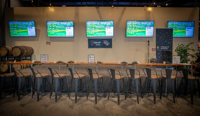 Long bar with metal stools and five wall-mounted monitors displaying a Windows desktop, barrels to the left, and banners for a College of Sciences event behind the counter.