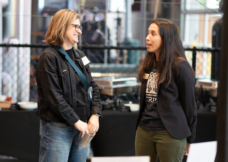 Two women talk and smile at a networking event, standing near a table with name badges visible in a modern venue behind them.