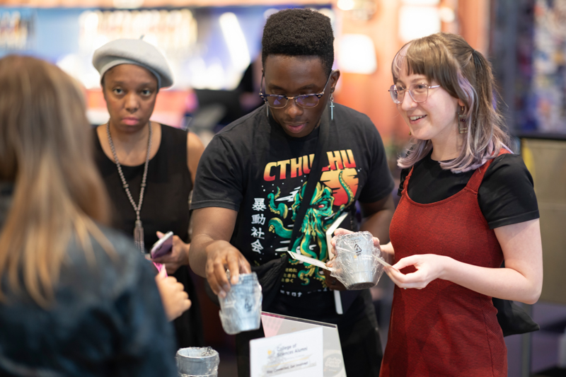 Three people at a booth share a moment as the man in the center hands a foil-wrapped cup to a woman on the right who smiles.