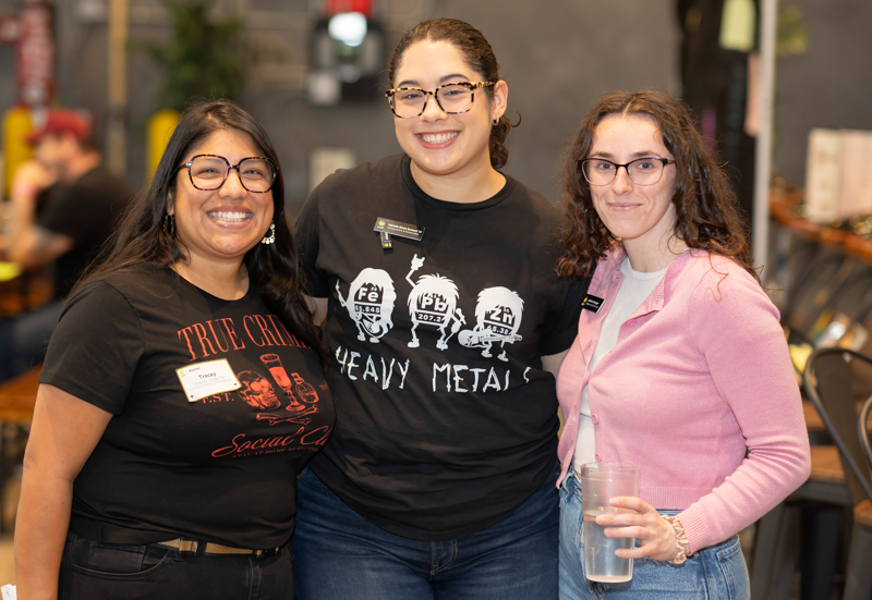 Three women posing together at an indoor event, smiling at the camera; two wearing graphic band shirts and name badges, the third holding a cup in a pink cardigan.