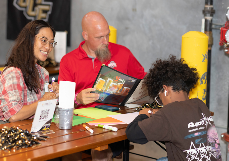 Three people sit at a wooden table in a workshop, looking at a glossy brochure together: a smiling woman on the left, a bearded man in a red polo in the middle, and a person with earbuds on the right writing/drawing.