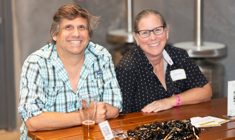Two smiling people sit at a wooden table with name badges at an indoor event, a beer glass in front of them.