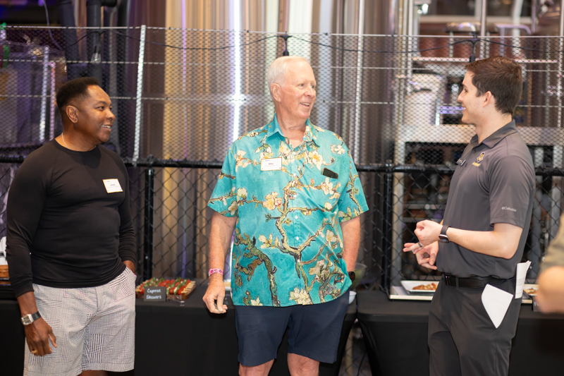 Three men chat smiling in a venue with a chain-link backdrop and snacks on a table behind them, name tags visible on their chests.