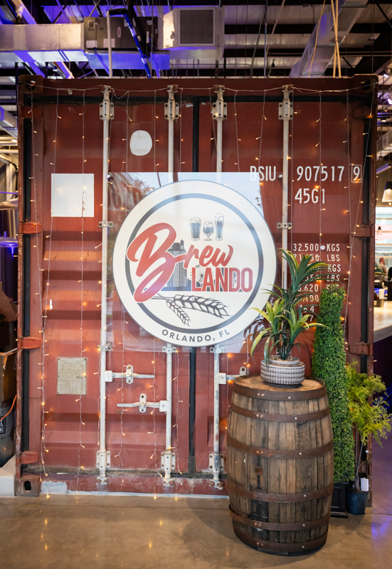 Shipping container booth with a circular 'Brew Orlando' logo sign, string lights, and a wooden barrel planter with a potted plant in front of it.