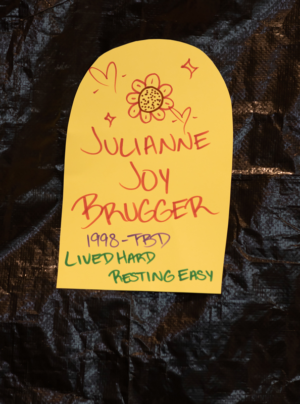 Yellow memorial tag with a red sunflower doodle and the name Julianne Joy Brugger; text reads 1998-? and 'Lived hard resting easy' in green.