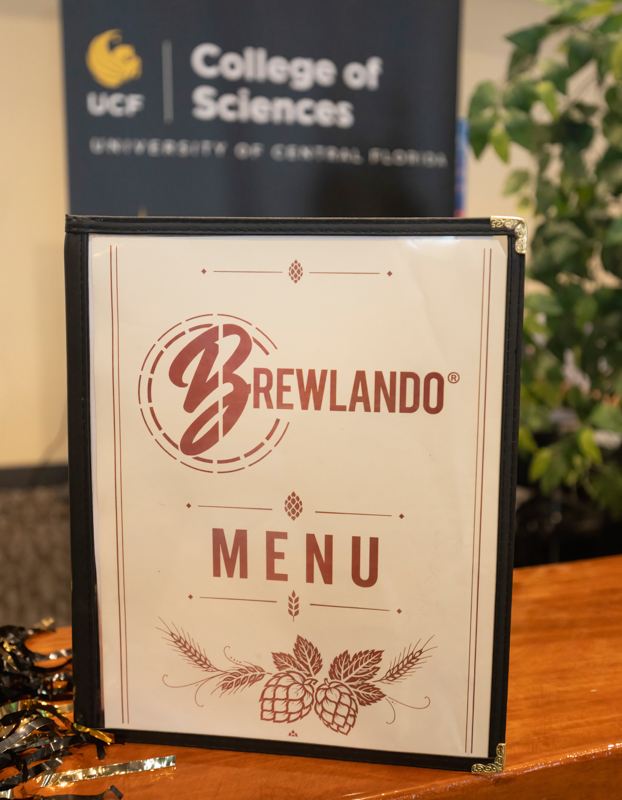 Menu binder displaying 'BREWLANDO MENU' with a hop graphic, on a wooden table; blurred 'UCF College of Sciences' sign in the background.
