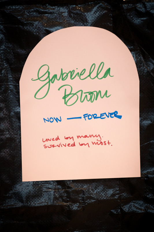 Pink tombstone-shaped card against a dark background, with green cursive text reading 'Gabriella Brown', blue 'NOW FOREVER', and red 'Loved by many, survived by most'.