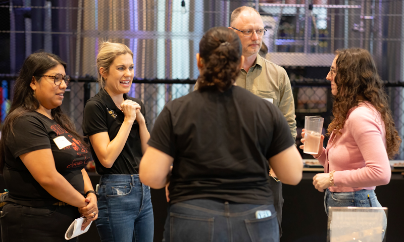 Five people standing in a casual group conversation at an event, with a woman in a pink sweater holding a cup and others smiling or listening.