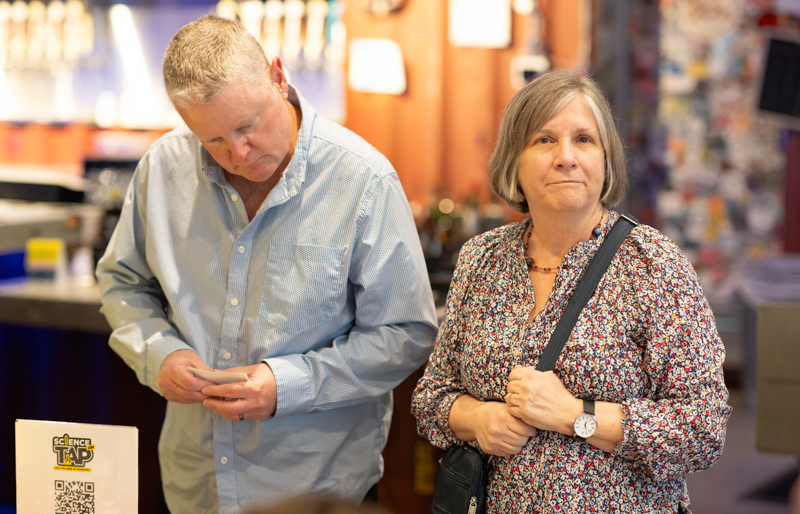 Man in a light blue shirt glances at his phone while a woman in a floral blouse stands beside him in a busy shop setting.