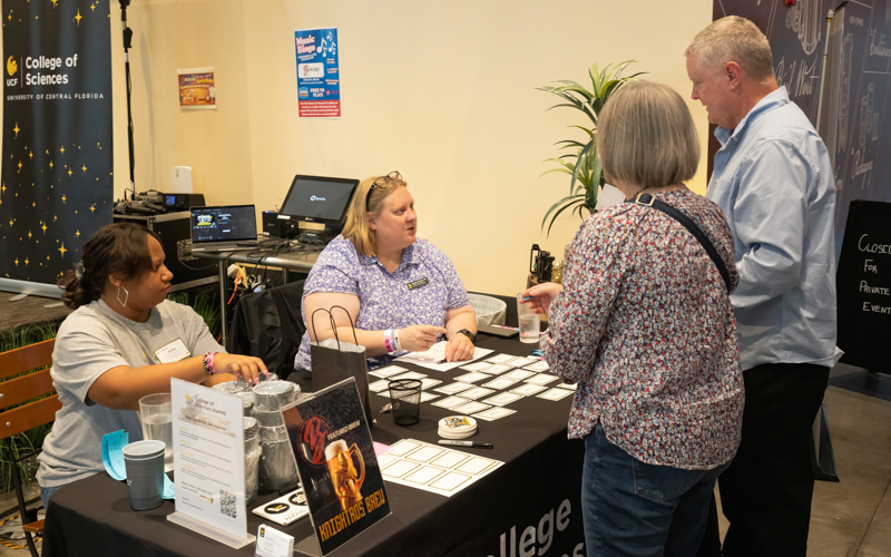 Staffed information booth for College of Sciences at an indoor event; two women seated behind a card-filled table as a couple talks with them.