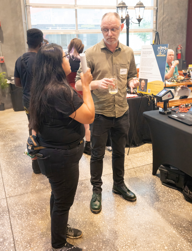 Man in a khaki shirt and glasses holds a glass while talking to a woman at a science event booth, with posters and equipment in the background.