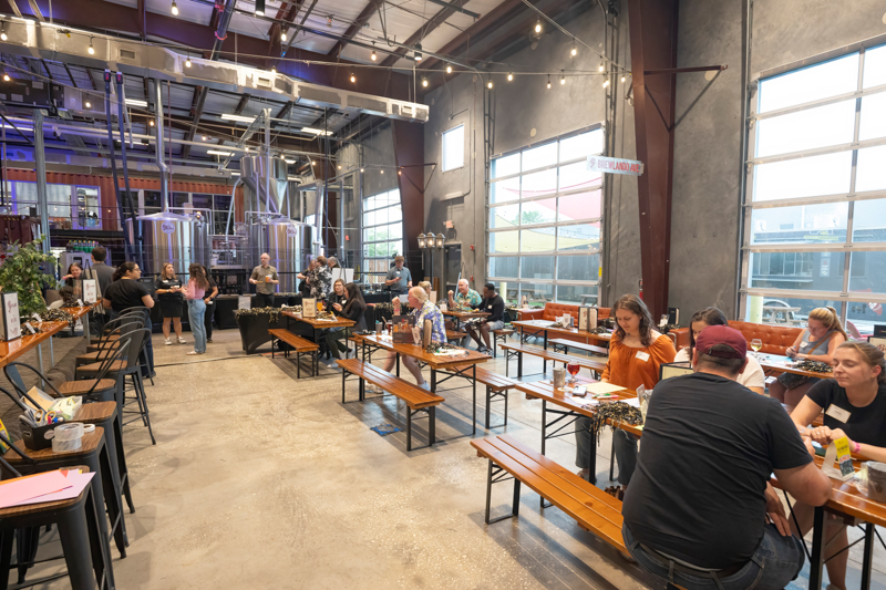 Industrial brewhouse event space with string lights where people sit at long wooden tables, enjoying a tasting or social gathering.