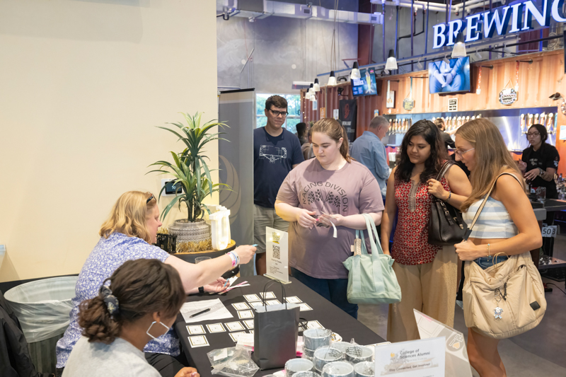 Group of young women examine a tabletop card display at a vendor booth, assisted by a staff member seated behind the table; brewery/bar area visible in the background.