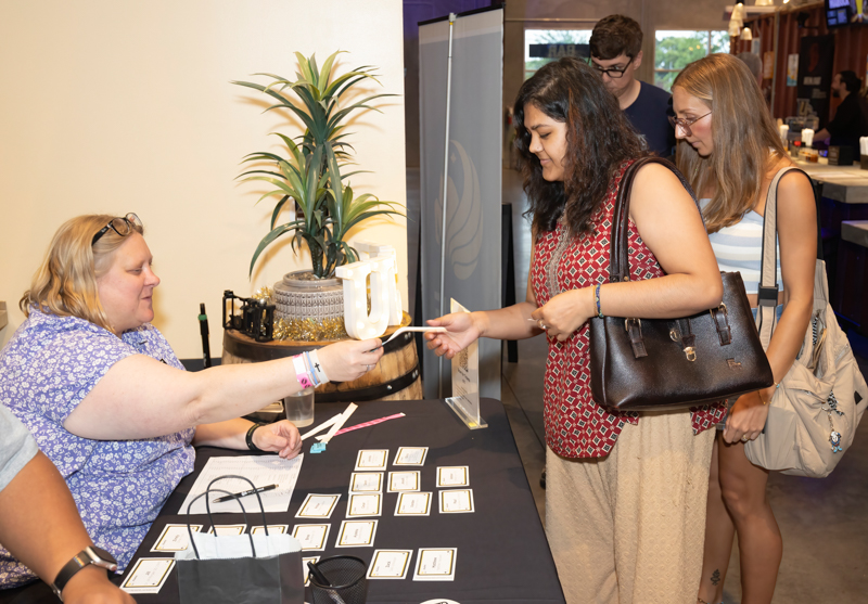 A woman at a booth hands a card to a visitor across a table with business cards laid out and a plant in the background.