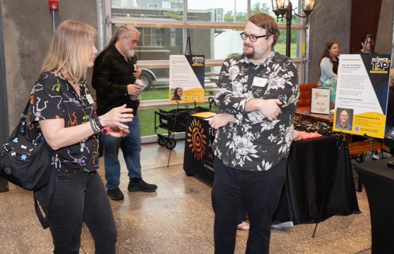 Two adults chat at an indoor event near a booth with posters and a table of materials in the background, a man in a black-and-white floral shirt facing a woman in dark clothing.