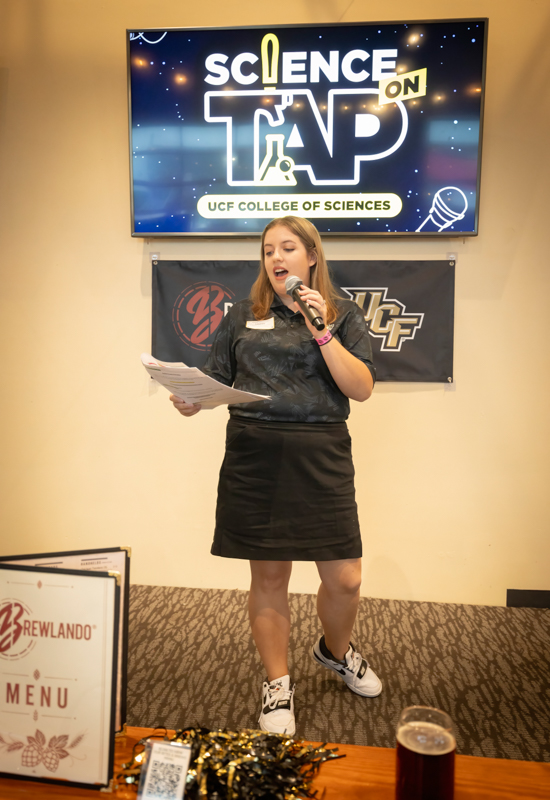 Woman presenting on stage at a Science on Tap event, holding notes and a microphone with the event logo on screen behind her.