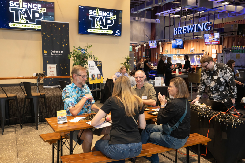 Group of five adults at a Science on Tap event, seated at a wooden table and chatting; a bar area and a 'Brewing' sign visible in the background.