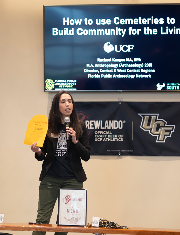A woman speaks into a microphone on a stage, holding a yellow notepad, with a large presentation screen behind her titled 'How to use Cemeteries to Build Community for the Living.'
