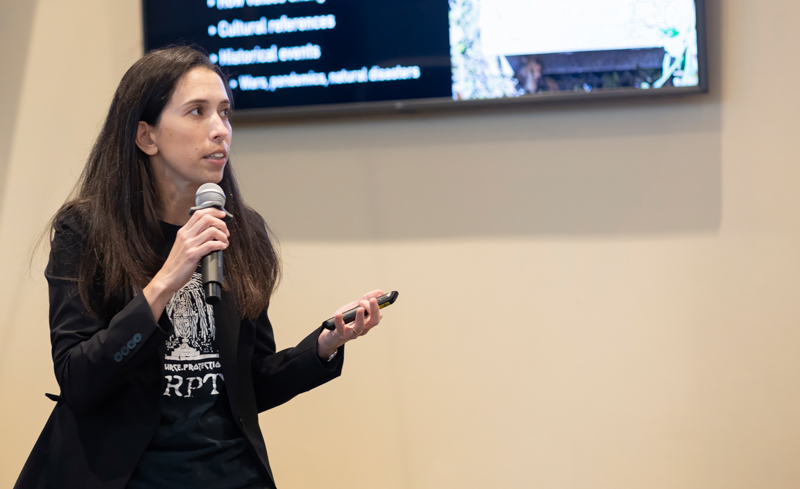 Woman presenting on stage, speaking into a microphone while holding a clicker with a large screen displaying slides behind her.