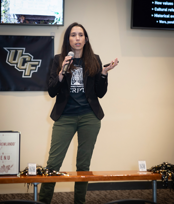 Woman in a blazer speaking on stage into a handheld microphone, gesturing with her other hand; UCF banner in the background.