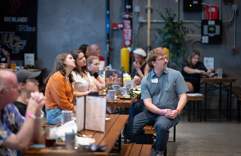 Group of adults seated at long wooden tables in a brewery venue, listening to a talk or event off-camera.