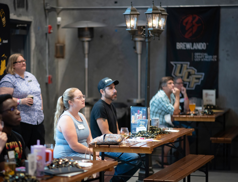 People seated at wooden tables in a brewery tasting room, listening to a presentation/brewery event.