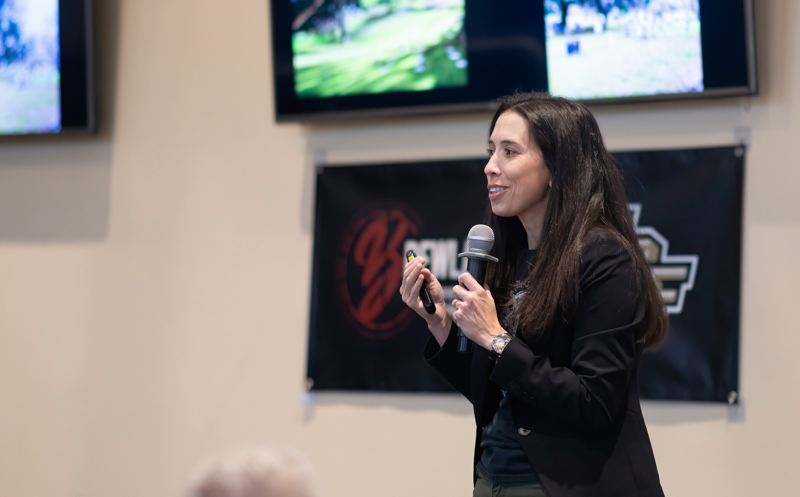 Woman in a blazer speaks into a handheld microphone while showing a clicker at a conference stage with banners in the background